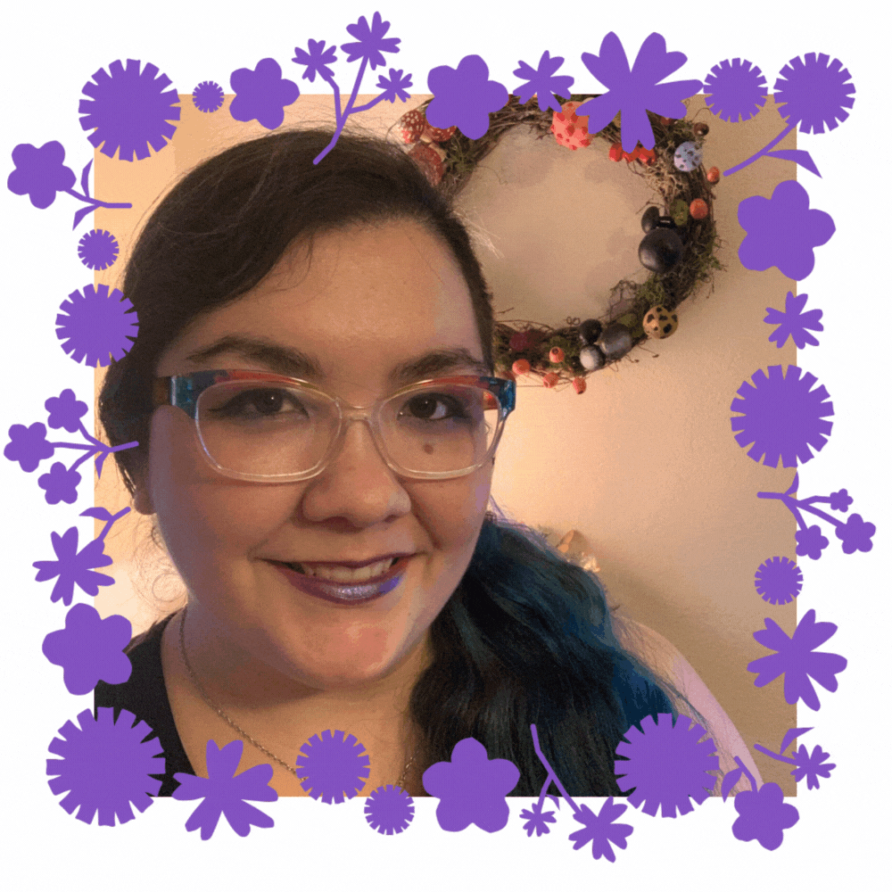 A woman with dark hair and rainbow glasses stands in front of a super cool mushroom wreath that he made. There are flowers that move slightly around the frame of the gif.