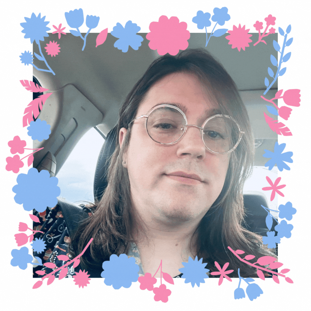 A picture of a trans person. They have brown hair and light skin. They wear round glasses and earrings. There are flowers that move slightly around the frame of the gif.
