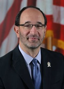 Tony Brancatelli, member of the Art House's board. Tony is wearing a black suit and blue tie in front of an American flag.