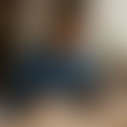 Man in denim shirt writing in notebook, using a laptop at a wooden desk. Background shows bookshelves and a potted plant. Focused mood.