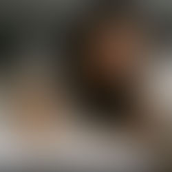 Woman in bed, looking tired with hand on forehead. Gray shirt, white sheets. Black alarm clock shows early morning time. Cozy setting.