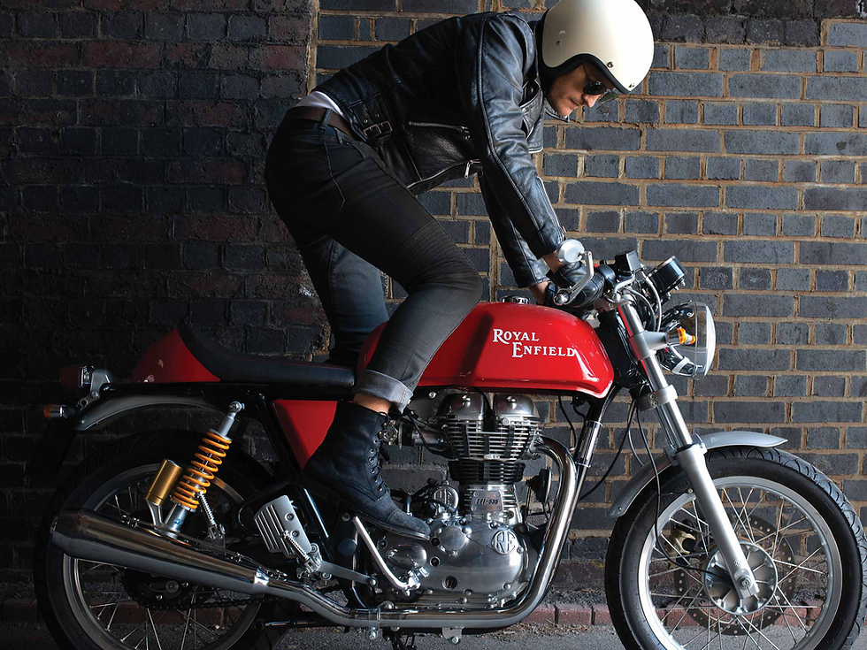 A rider in a black leather jacket and cream helmet leans over a vibrant red Royal Enfield Continental GT 650, parked against a dark brick wall. The bike’s classic cafe racer design is highlighted by its chrome engine, orange rear shocks, and the "Royal Enfield" logo on the fuel tank.