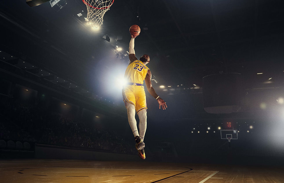 LeBron James, in his iconic Lakers #23 yellow jersey, soars through the air for a powerful dunk under bright arena lights. He’s mid-air, right hand extended towards the hoop with the ball, showcasing peak athleticism against a dark, hazy background with a visible crowd and scoreboard.
