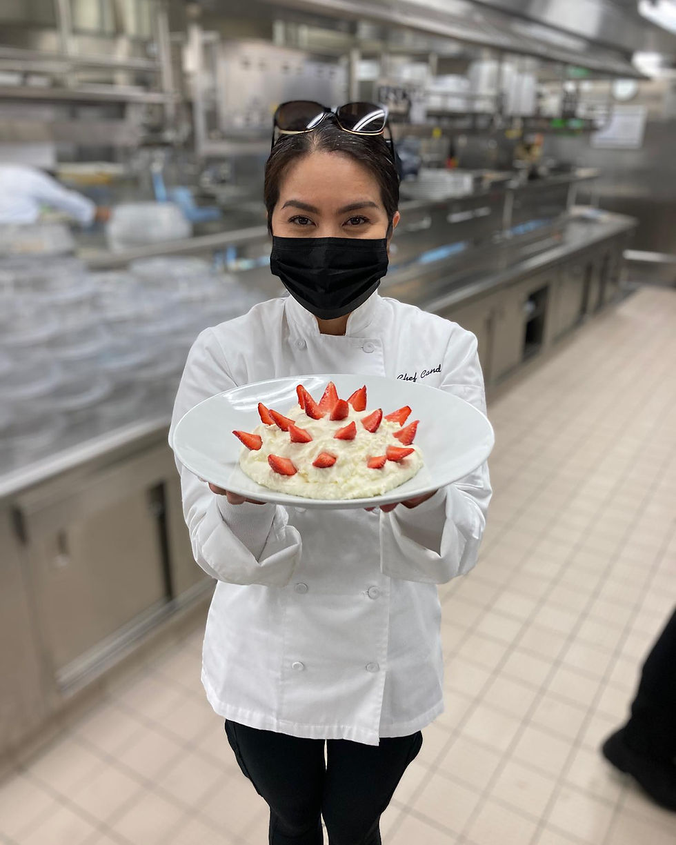 Chef in a white coat presents a dessert topped with sliced strawberries in a commercial kitchen. She wears a black mask and sunglasses on her head.
