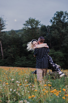 blurry engagement photo of a blond girl in a black floral dress and a man in a black stripped shirt in a flower field with a full moon