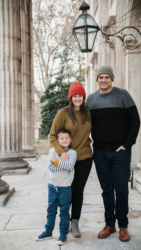 Family of three wearing winter hats standing in front of columns in Old City Philadelphia in front of first national bank of the united states