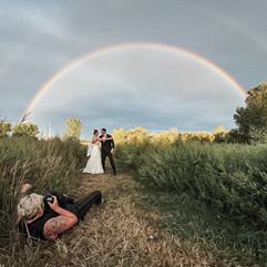 Gabby Smarr laying on the ground photographing a bride and groom under a double rainbow in a field in front of the philadelphia skyline
