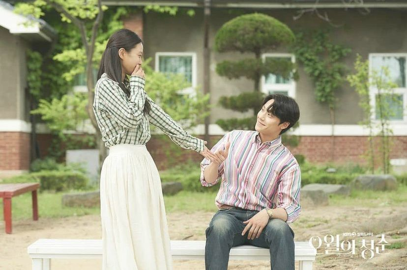 Woman holding hands with a seated man in a sunny courtyard in Kdrama Youth of May