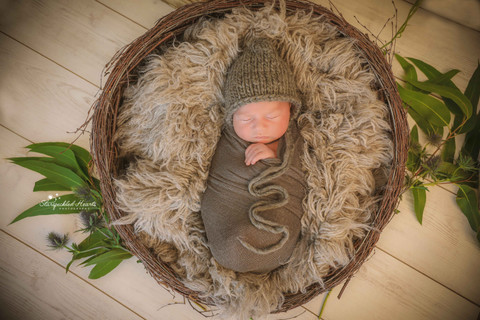 sleeping baby boy lying in a round wicker basket stuffed with a grey fluffy rug, swaddled and wearing a bonnet