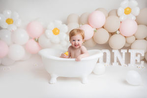 an adorable daisy themed cake smash setup for a little girl, featuring the colours beige and pink along with daisy balloons. the birthday girl is sat in a white rolltop bath, smiling