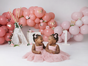 Twin girls in pink dresses smile at each other, surrounded by pink balloons, flowers, and a small teepee. A large white "1" stands nearby.