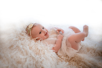 baby girl wearing a white lacy romper, lying on a white fluffy rug