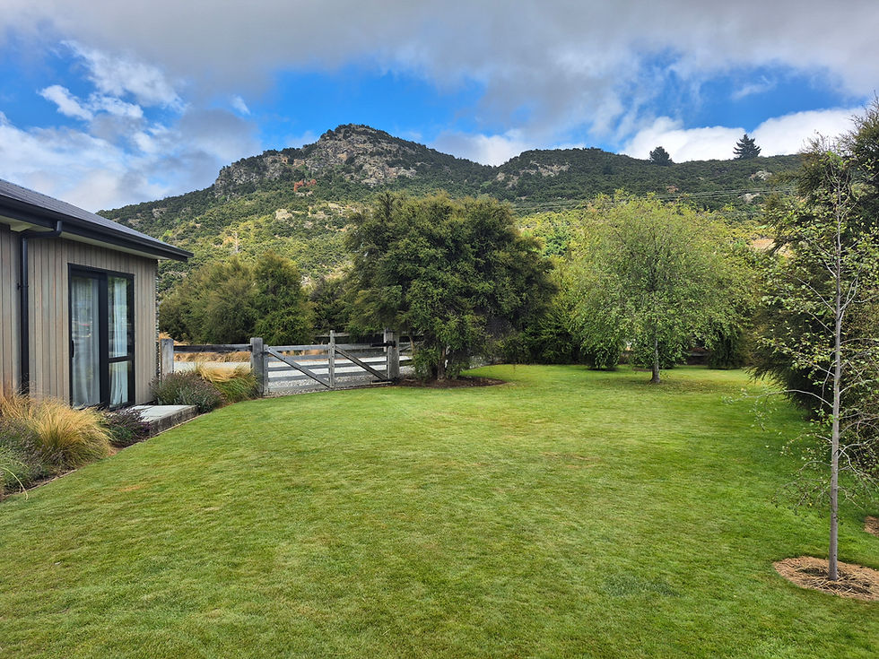 House exterior with large green lawn, surrounded by trees and mountains. A wooden fence and gate lead to a gravel path. Bright, cloudy sky.