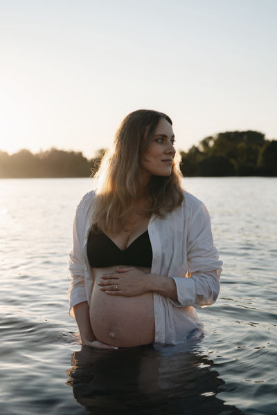 Frau während Schwangerschaft steht im Wasser bei Sonnenuntergang.
