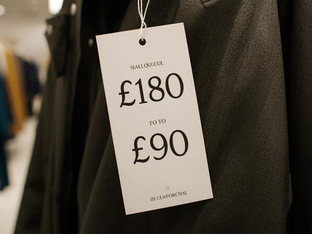 Price tag showing a sale from £180 to £90 on dark fabric in a clothing store. Background has blurred racks of colourful clothing.