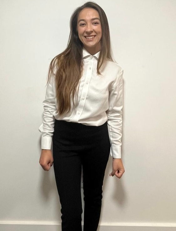 Smiling woman in white shirt and black pants stands against a plain white wall. Her long hair is loose, creating a cheerful, professional vibe.