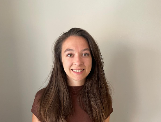 Smiling woman with long brown hair in a brown shirt stands against a plain beige wall.