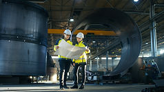 Male and Female Industrial Engineers Look at Project Blueprints While Standing Surround By