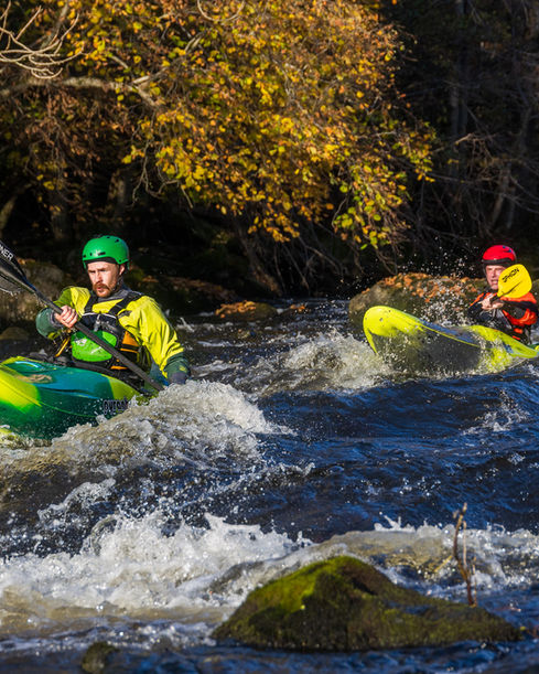 Två paddlare i färgglada kajaker paddlar tillsammans genom forsande vatten i höstmiljö.