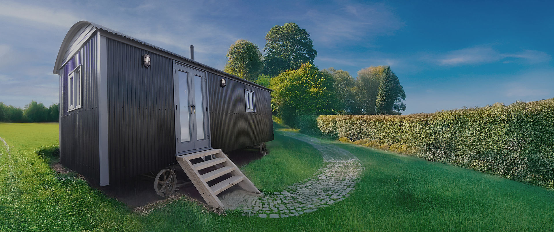 Shepherd’s hut with steps and wheels, set beside a cobbled path in a peaceful UK countryside location, surrounded by green hedges and trees.