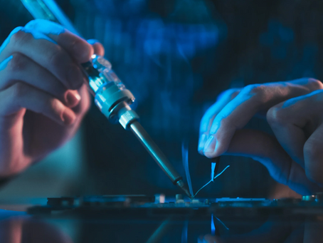 Hands soldering electronic components in low light. Blue tint creates a focused, technical mood, with visible smoke and circuit details.