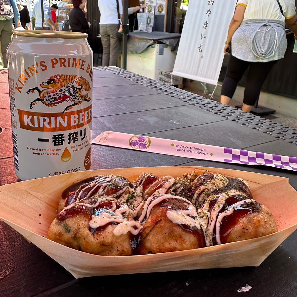 Kirin Beer can beside sauced takoyaki at an outdoor Japanese food stall, with vendors and Japanese text in the background.