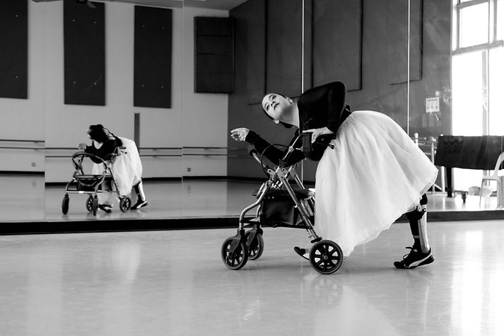 A black and white photograph of Vanessa dancing with her rollator (walker). She is leaning onto her walker with one arm gently leaning on the handle and the other arm leaning on the backrest. Her serene face is gently looking out into the distance. Her legs are positioned in a mid-walking stance. One of her leg braces is visible. Her hair is pulled back into a ballet bun. She is wearing a tulle dress; dark top, light and fluffy skirt. She is in a well lit dance studio where mirrors are visible and her reflection is present.