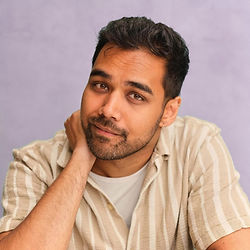 A headshot of Mohit Mathur, who has short dark brown hair and brown eyes. Mohit is wearing a light beige striped shirt over a white T-shirt, and is resting their head on one hand and looking directly at the camera with a gentle smile against a soft purple background.