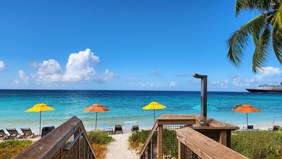 View from the boardwalk steps down to the Lighthouse Point family beach with orange and yellow umbrellas, turquoise water, and the Disney cruise ship in the distance