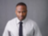 Man in a light shirt and dark tie stands against a gray backdrop, looking confidently at the camera. Mood is serious and professional.