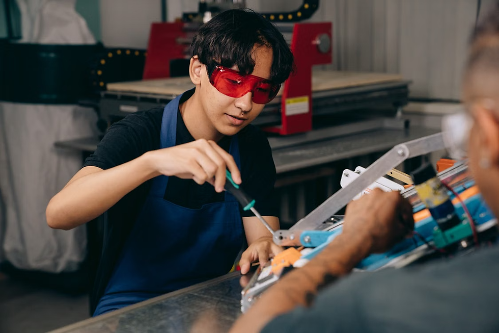 Young man in red safety glasses and blue apron uses a screwdriver on a machine in a workshop, with another person nearby.