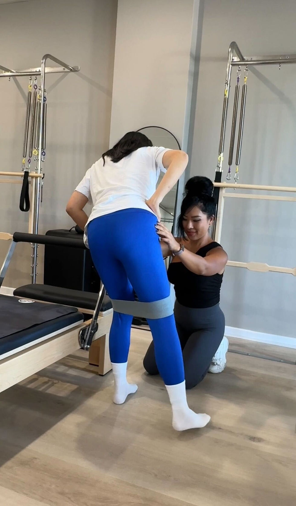 Pilates instructor kneels to steady a woman in blue leggings using a resistance band in a bright studio, focused and supportive