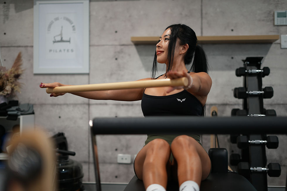 Woman doing Pilates with a wooden pole, seated on a gym bench. Background has gym equipment and a "Pilates" poster. Calm expression.