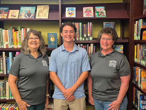 Two adult women wearing shirts with the Friends of the Library logo standing with a teenage boy wearing a polo shirt and glasses. They are all standing in front of a book shelf.