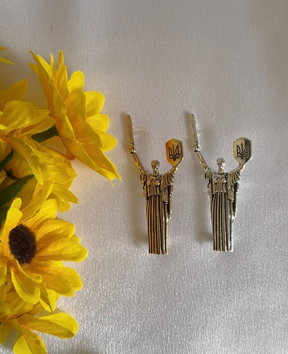 Thumbnail: two motherland monument brooches, silver and bronze gently placed on the white cloth close to sunflowers
