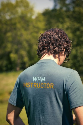 A medium close-up from behind a person with dark, curly hair wearing a blue-grey t-shirt. The back of the shirt features text reading "WHM" in light blue and "INSTRUCTOR" in yellow. The background shows a softly blurred green field and trees under natural sunlight.