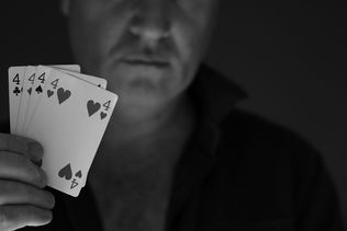 A black and white close-up shot of a man's hand holding a winning poker hand of four number four cards, with his blurred face in the background.