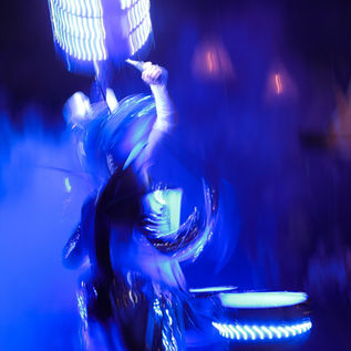 An abstract, motion-blurred photograph captures a drummer performing on an LED-lit drum kit. Saturated blue and violet lighting dominates the scene, with glowing drums and costume reflecting the light into sweeping trails. The performer's face is obscured by motion, but raised white-gloved hands gripping drumsticks are visible, emphasising the speed, rhythm, and excitement of the performance.