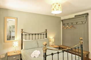 A brightly lit cottage bedroom featuring a double bed with a black and brass metal frame, made with green-and-white checkered bedding and three rolled white towels at the foot. Sage green walls frame the room, adorned with a vertical mirror on the left reflecting a bunk bed, and a wall-mounted wooden coat rack with hangers on the right above a matching bench. Matching lamps with patterned shades sit on nightstands on either side of the bed, complementing a central cylinder pendant light overhead.