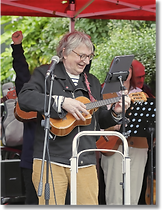 A smiling musician with grey hair and glasses plays a ukulele under a red canopy, looking at lyrics on a digital tablet during a live group performance.