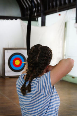 RA medium close-up from behind a person with dark brown hair styled in two French braids, wearing a blue-and-white striped t-shirt. The person is aiming a black bow, with their right arm pulled back, targeting a square, multi-colored archery target with concentric rings of blue, red, and yellow in the blurred background. The indoor setting features a wooden floor and white walls.