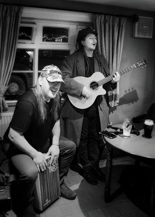A black and white photograph of two musicians performing inside a pub, with one singing and playing an acoustic guitar while the other plays a cajón drum.