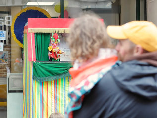 A traditional Punch and Judy style puppet theater booth on a street, with spectators watching from the foreground.