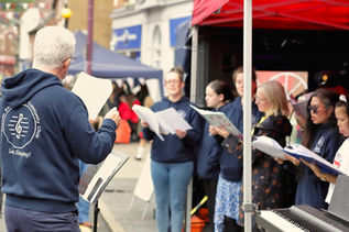 An outdoor performance scene showing a community choir. In the foreground on the left, a choir director with short grey hair is seen from behind. He wears glasses and a navy blue hoodie with a circular logo on the back reading "Community Choir" around a treble clef symbol. He holds a piece of sheet music in his left hand and faces his choir.