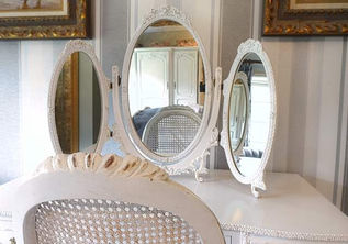 An eye-level shot focusing on an elegant, vintage-style vanity setup. A white, tri-fold tabletop mirror with detailed, ornate carvings along its oval frames sits at the back of a smooth white dressing table. The mirrors reflect parts of the room, including a white wardrobe and the woven cane back of a matching chair positioned in the foreground. The background features light gray and white vertically striped wallpaper flanked by two gold-framed paintings on either side.