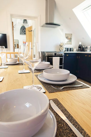 A bright, low-angle perspective looking down a long wooden dining table set for a meal. Each place setting features a textured dark placemat, a light grey plate and matching bowl stack, silverware, and a tall, clear wine glass. The background opens up to a modern kitchen with dark blue cabinets, a stainless steel stove, a range hood, and a slanted ceiling with a skylight.