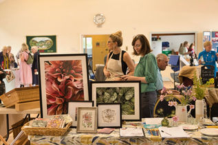 Inside a bright community hall, visitors are gathered around a stall displaying botanical artwork and handmade items. In the foreground, a table is covered with framed floral and leaf prints in a range of sizes, alongside baskets of greetings cards, notebooks, pencils, and small decorative vases containing flowers. Two women stand behind the display, looking at the artwork and talking, while other visitors browse nearby stalls in the background. Additional exhibitors and displays can be seen across the hall, creating a busy, welcoming atmosphere typical of a local arts and crafts fair. The warm lighting and open space give the event a friendly community feel.