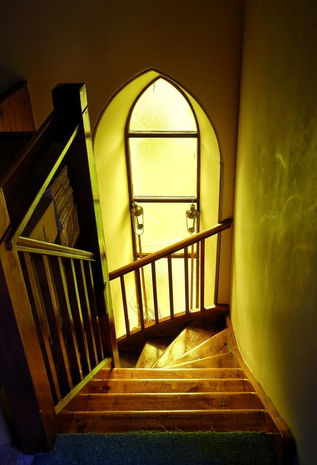 A high-angle perspective looking down a winding wooden staircase illuminated by a warm, golden glow. The polished wooden steps curve downward around a dark wooden banister with vertical slats on the left. In the center background, a tall, arched Gothic-style window with frosted glass panels allows bright, yellow-tinted light to flood the stairwell, casting soft reflections on the stairs and the textured yellow wall on the right. Two small, decorative metal lanterns hang symmetrically on either side of the lower window frame.