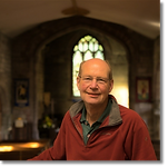 A portrait of a smiling, middle-aged man with glasses and a red pullover, standing inside a dimly lit historic church with an arched window behind him.