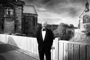 A black and white portrait of a smiling, bearded man in a tuxedo standing on a bridge with a historic stone castle and chapel in the background.
